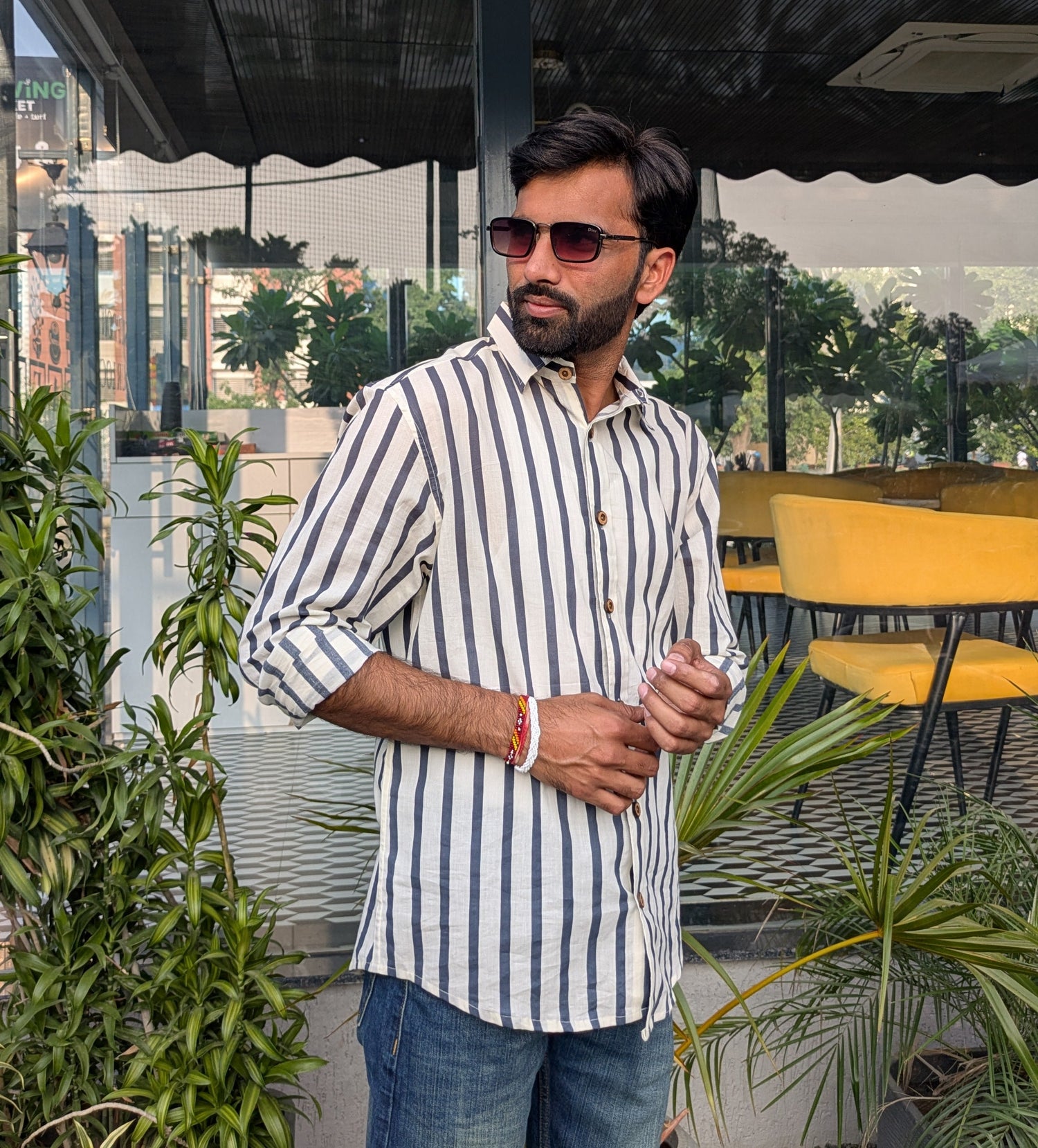 Man wearing a striped shirt and sunglasses standing outdoors with plants and furniture in the background